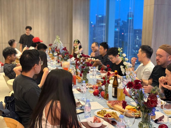 A group of people are gathered around a long dining table set with elaborate floral centerpieces, food, and drinks, with a city skyline visible through the windows in the evening.