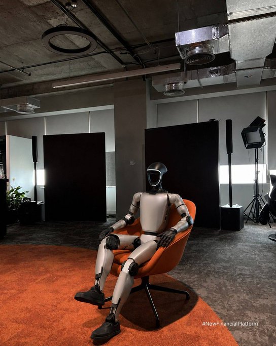 A humanoid robot sits in an orange chair in a modern office studio setting, with the hashtag '#NewFinancialPlatform' visible in the corner.