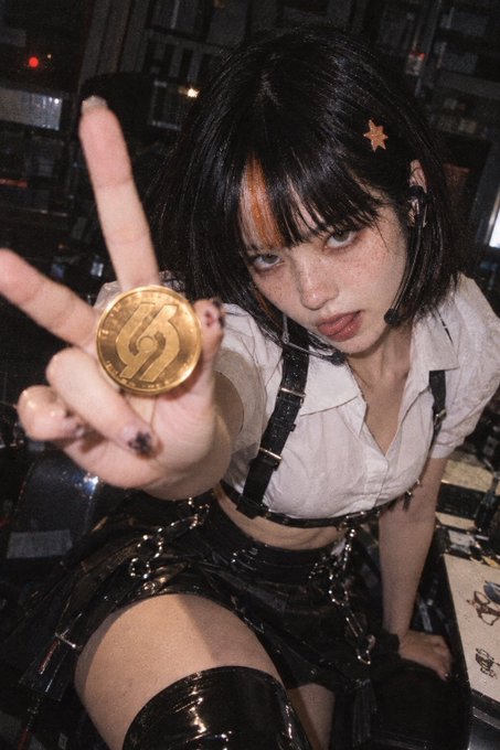 A close-up photograph of a young woman with short dark hair and an orange star hairclip, wearing a white shirt with black leather harness straps, posing while holding up a gold coin with a stylized "S" logo and making a peace sign.