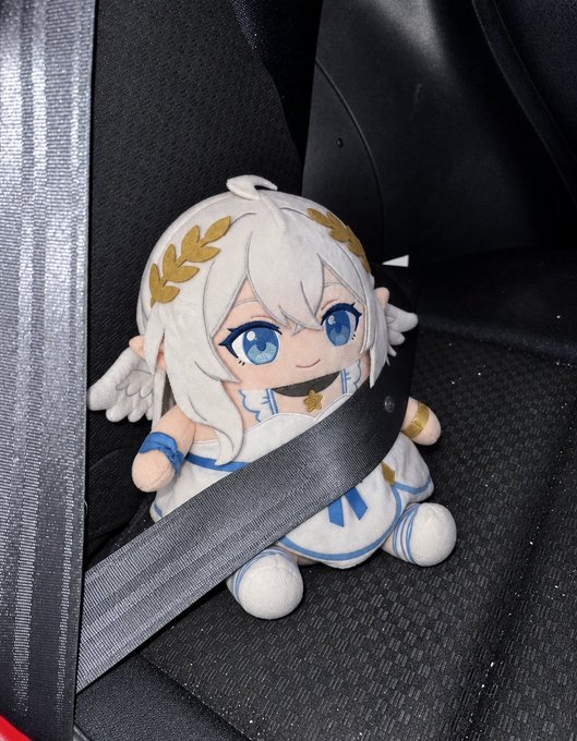 A plush toy of a white-haired anime character with blue eyes, a golden laurel wreath crown, and small wings is secured by a seatbelt on a car seat.