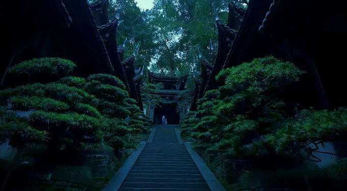 A moody photograph of a person in white walking up stone stairs toward a traditional Asian temple gate, flanked by lush green bonsai-style trees.