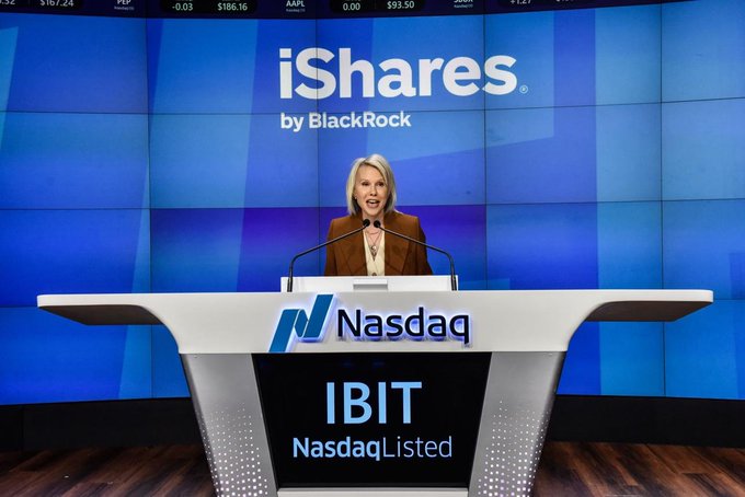 A photograph of a woman speaking at the Nasdaq podium, with the iShares by BlackRock logo displayed on the screen behind her and 'IBIT NasdaqListed' on the podium.