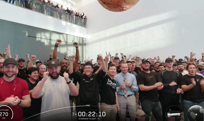 A large group of people cheers inside a building during what appears to be a SpaceX Starship launch event, with an overlay showing 'T+ 00:22:10 STARSHIP FLIGHT 14' and altitude data.