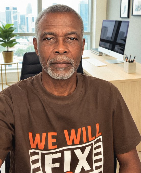 An older man with a gray beard sits in an office setting wearing a brown t-shirt that reads 'WE WILL FIX IT' in orange and white text.