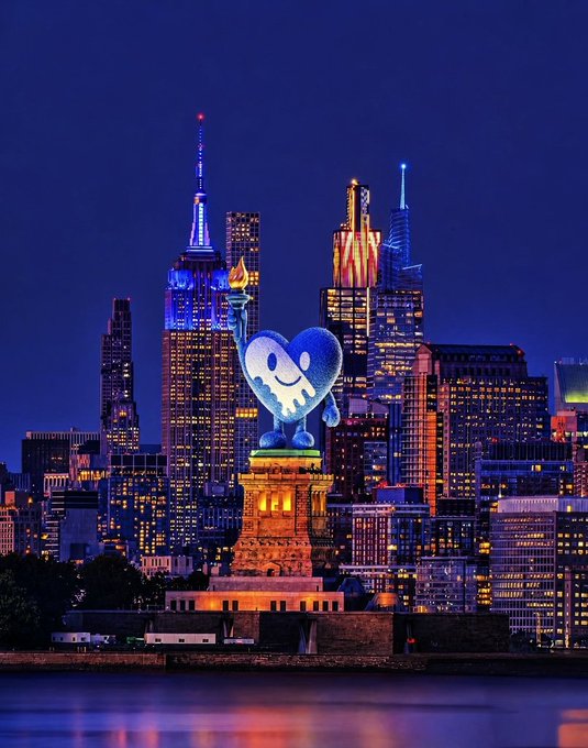 A nighttime cityscape of New York City featuring the Statue of Liberty, with a large glowing blue heart-shaped emoji character projected onto the pedestal, and the Empire State Building lit in blue in the background.
