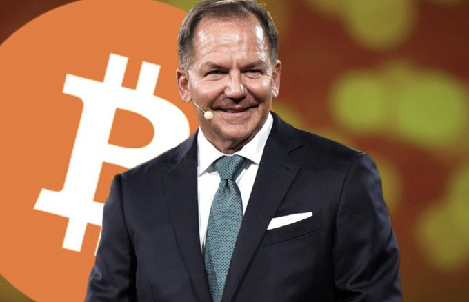 The image shows billionaire hedge fund manager Paul Tudor Jones speaking at an event, with a large Bitcoin logo displayed prominently behind him. His firm's reported $22.9 million investment in BlackRock's spot Bitcoin ETF (IBIT) aligns with his well-known history of endorsing Bitcoin as an inflation hedge and store of value.