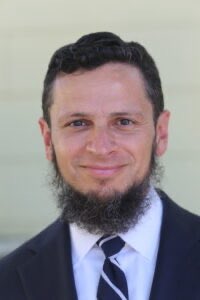 A professional headshot of a bearded man in a dark suit, white shirt, and striped tie smiling at the camera.