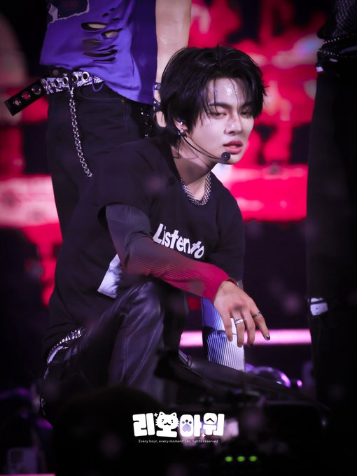 A performer with dark hair, wearing a headset microphone and a black long-sleeve shirt reading "Listenvio," crouches on stage during a concert, with Korean text at the bottom reading "리얼타임" (Real-time). The image aligns with the post's caption referencing KM Chart Award 2026, highlighting the artist's stage presence during the event.