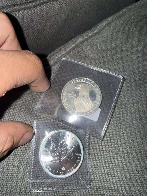 A hand holds two silver bullion coins in clear plastic protective sleeves; the top coin features a Native American chief with 'UNITED STATES OF AMERICA' text, while the bottom coin displays a maple leaf design with 'CANADA' and 'FINE SILVER 1 OZ ARGENT PUR' inscriptions.