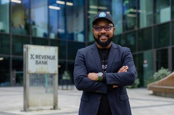 A man wearing a dark blazer, black t-shirt, glasses, and a dark cap stands with arms crossed smiling in front of a building with a sign reading 'X REVENUE BANK'.