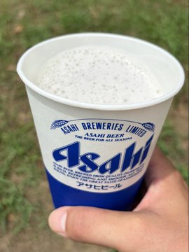 A hand holding a plastic cup of Asahi Super Dry beer with Japanese text 'アサヒビール' (Asahi Beer), photographed outdoors against grass.