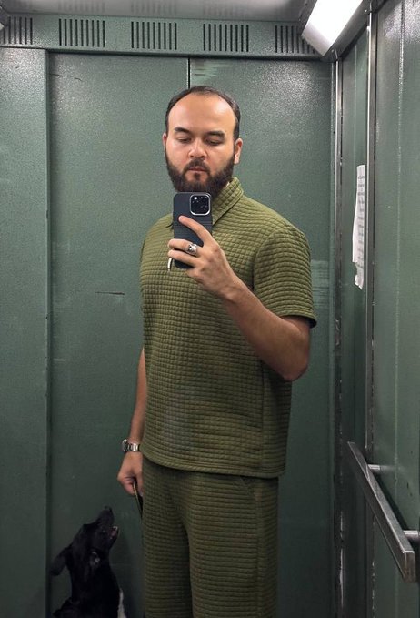 The image shows a man standing in an elevator holding his cell phone up to take a selfie. A dog is present close by, possibly waiting for someone or accompanying them in their ride. The caption of the photo indicates that they have posted the selfie on social media and it has gained popularity reaching $1.8M.
