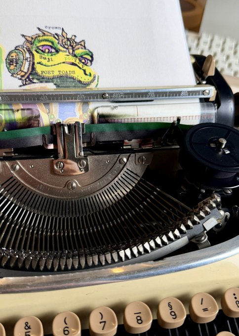 A close-up of a vintage typewriter with paper loaded, featuring a hand-drawn or printed image of a 'POET TOADS' character at the top of the page.