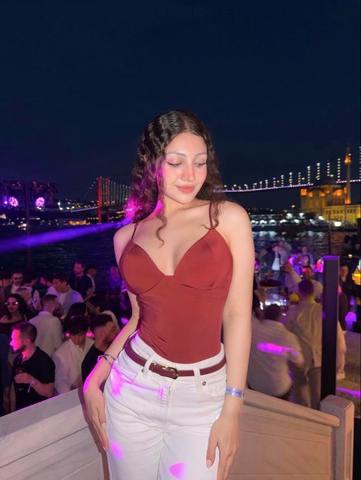 A 21-year-old poses in a burgundy top and white jeans at a vibrant waterfront nightclub, with an illuminated bridge and crowded party scene in the background capturing the energy of a milestone birthday celebration.