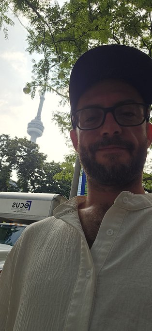 A selfie of a bearded man wearing a black cap, glasses, and a light-colored button-up shirt, with a tall observation tower visible in the background through tree foliage.