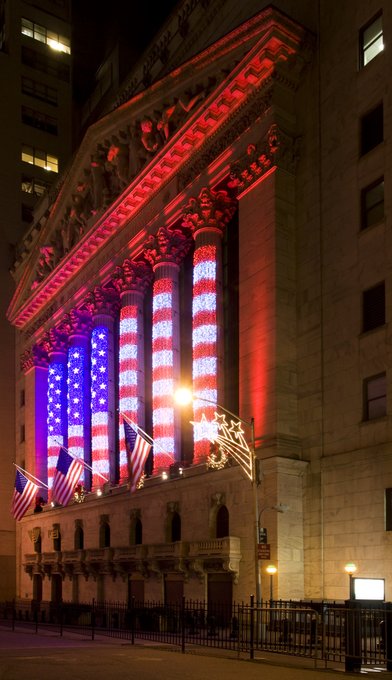 The New York Stock Exchange building at night, illuminated in red, white, and blue lights forming the American flag on its columns.