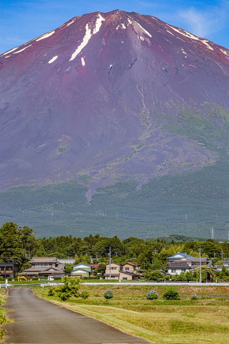 A scenic landscape view shows a small village with houses and green fields in the foreground, with the large, snow-streaked Mount Fuji rising prominently in the background under a blue sky.