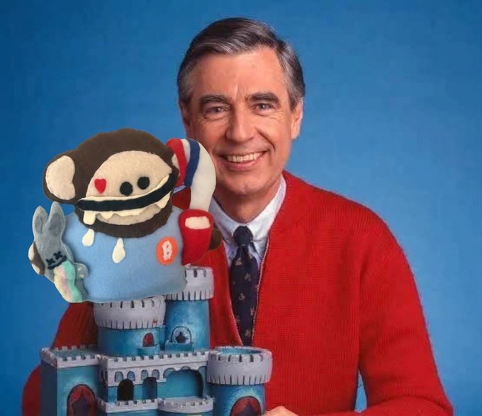 Fred Rogers in his iconic red cardigan smiling next to a plush puppet wearing a blue shirt with a Bitcoin 'B' logo, alongside a small blue castle with a Bitcoin 'B' on one of its turrets.