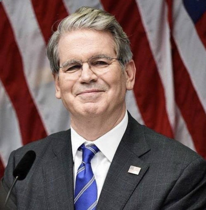 A man with gray hair and glasses wearing a dark suit, blue striped tie, and American flag pin stands at a microphone in front of an American flag backdrop.