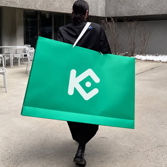 A person in black attire walks away carrying an oversized green KuCoin-branded shopping bag with the distinctive white "K" logo prominently displayed on its side. The image plays on the cryptocurrency slang phrase "securing the bag," visually representing a triumphant KuCoin user showing off their crypto gains through the larger-than-life branded bag.