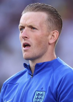 A close-up of England goalkeeper Jordan Pickford wearing a blue England national team training jacket, looking focused with his mouth slightly open. The image relates to the post about the Everton shot-stopper's reported reckless driving incident in his luxury Lamborghini.