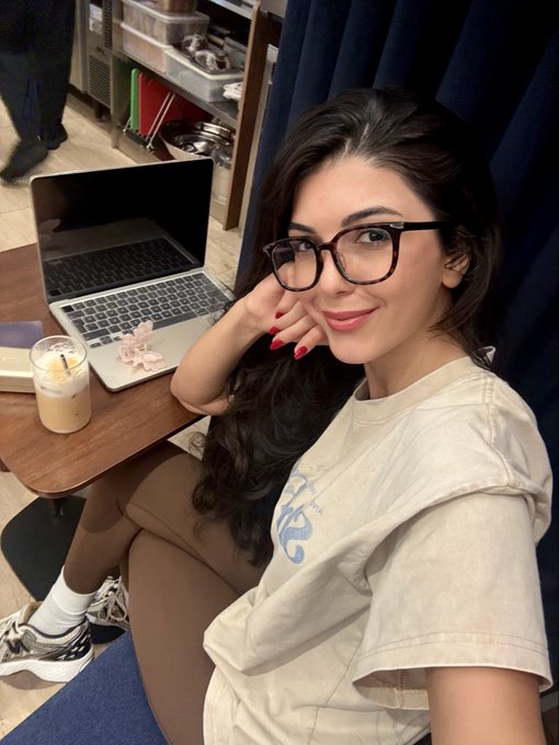 A young woman with long dark hair and black glasses smiles from a workspace, leaning casually next to a laptop with a candle and coffee on the desk, embodying a cozy "gm from Singapore" morning vibe.