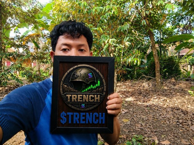 A young man in a blue long-sleeved shirt holds up a framed artwork featuring a soldier helmet emblem and the text 'TRENCH' and '$TRENCH' against a backdrop of lush green foliage.