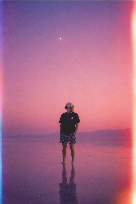 The image features a man standing alone at the edge of an ocean, wearing sunglasses and shorts. He appears to be looking out over the water or possibly contemplating something. The photo has been edited with purple and orange hues, giving it a dreamy atmosphere. The caption "GM bored ape yacht club" suggests that this scene might have been staged for a promotional purpose related to a yacht club or a similar establishment.