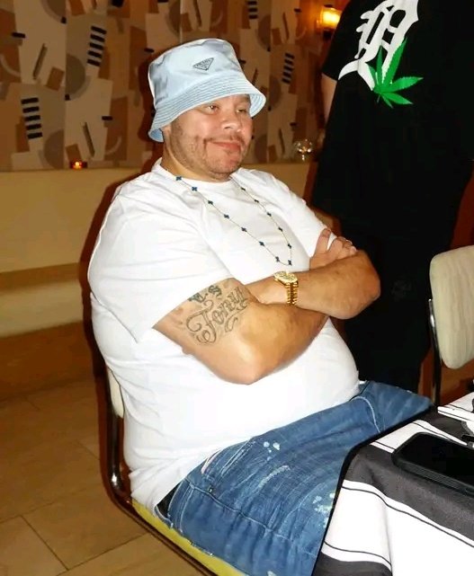 The image features a man wearing a hat sitting at a table with his arms crossed. The background of the photo shows another person standing behind him. There are two chairs visible in the scene - one where the man is seated, and another chair that appears to be empty. A dining table can also be seen in the background.