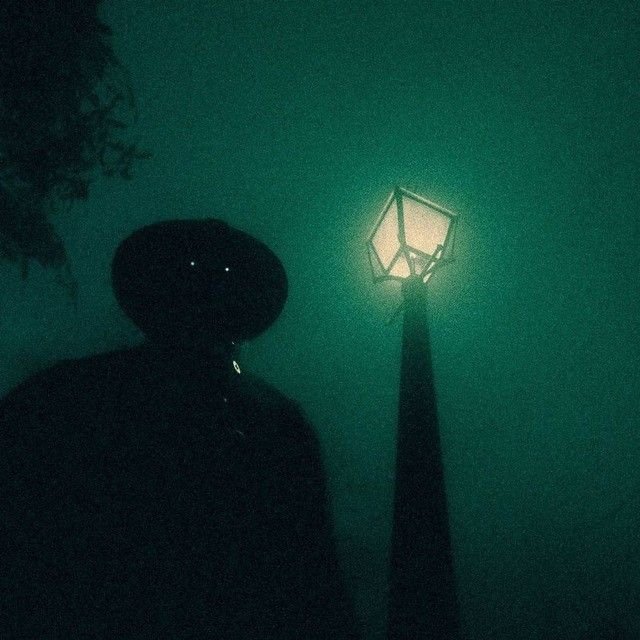 A dark, atmospheric image of a silhouetted figure wearing a wide-brimmed hat standing next to a glowing vintage street lamp enveloped in green mist.