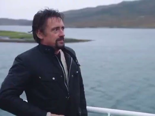 A man with a beard wearing a dark jacket stands near a body of water with land visible in the background.