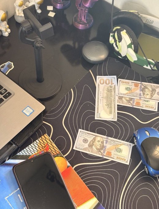 A messy desk setup featuring a laptop, headphones, a microphone, a phone, a computer mouse, and several hundred dollar bills scattered on a black and white patterned mousepad.