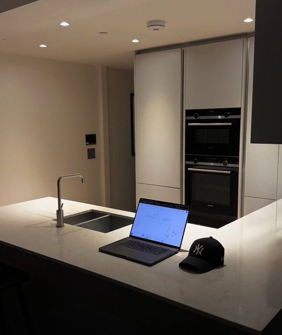 A modern kitchen features a white marble island with an open laptop and a black New York Yankees 'NY' baseball cap, alongside built-in ovens and a sink.