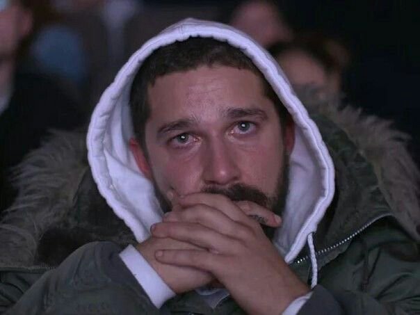 A close-up shot of Shia LaBeouf wearing a gray hooded jacket, staring intensely ahead with his hands clasped over his mouth.