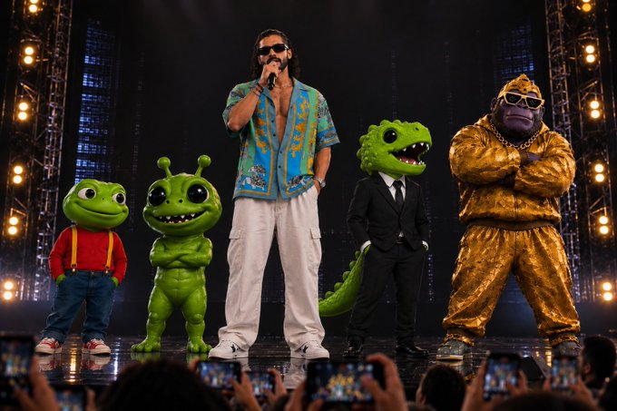 A concert stage scene featuring a male performer with sunglasses, a colorful shirt, and white pants standing center, flanked by costumed characters including green aliens, a dinosaur in a suit, and a figure in a gold outfit, with an audience filming on smartphones in the foreground.