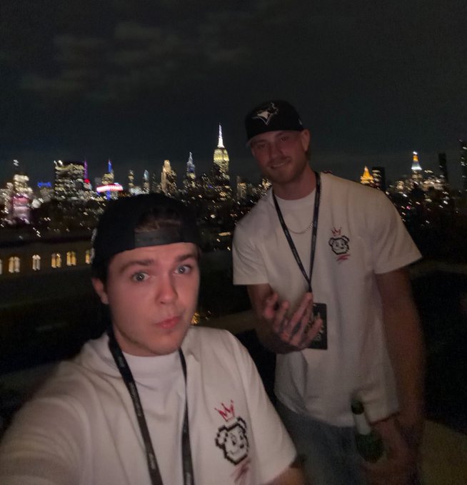 Two men taking a selfie at night with the New York City skyline illuminated in the background; one wears a white t-shirt with a crown logo and lanyard, while the other wears a black cap and holds a bottle.