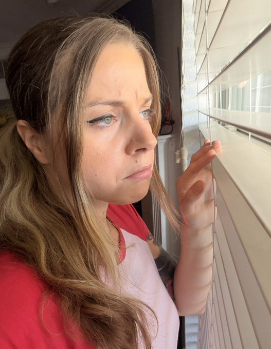 A close-up selfie of a woman with brown hair looking out a window with white blinds, wearing a red shirt over a white top.