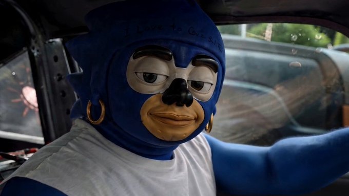 A person wearing a blue Sonic the Hedgehog mask with 'I Love to Cook' written on it sits inside a vehicle, with a tired-looking mask face and gold hoop earrings.