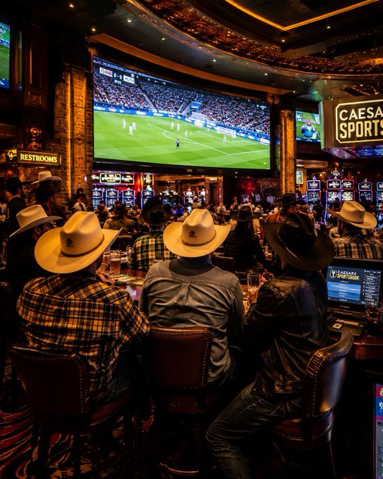 A crowded sportsbook interior, likely at a Caesars Sportsbook location, with patrons wearing cowboy hats watching a soccer match on a large screen, with slot machines and a 'RESTROOMS' sign visible in the background.