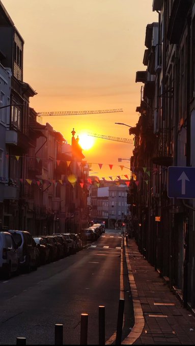 A sunset glows between buildings at the end of a narrow city street decorated with colorful pennant flags.