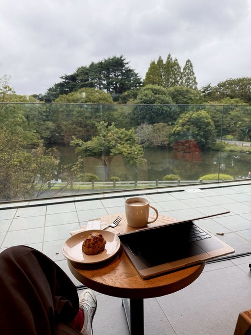 A relaxing cafe scene featuring a small round table with a laptop, a cup of coffee, and a pastry, set against a glass window overlooking a lush green park with a pond.