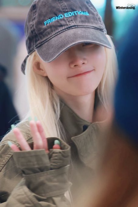 A young woman with blonde hair wearing a dark gray 'FRIEND EDITIONS' cap and a green jacket, smiling slightly while gesturing with her fingers, with a 'Winterclosi̇ng' watermark in the corner.