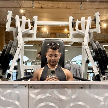 A man takes a mirror selfie while seated on a gym machine, likely capturing his first day at the Colosseum accelerator in San Francisco by combining his pitch prep with a chest workout. The image visually reinforces his "pitch reps between chest reps" theme, blending startup hustle with fitness motivation.