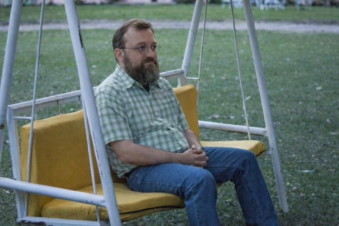 A bearded man wearing glasses and a green checkered short-sleeve shirt sits on a yellow-cushioned swing outdoors on a grassy area.