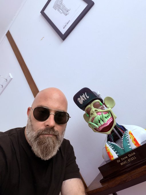 A bald man with a gray beard and sunglasses poses for a selfie next to a grotesque green 'Dr Dave Coin' sculpture wearing a hat with 'BAYC' written on it, dated WTF #211.