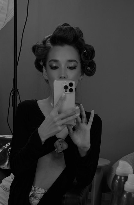 A black and white photo of a woman taking a mirror selfie with her phone, wearing hair rollers and a black top, showing a ring on her finger.