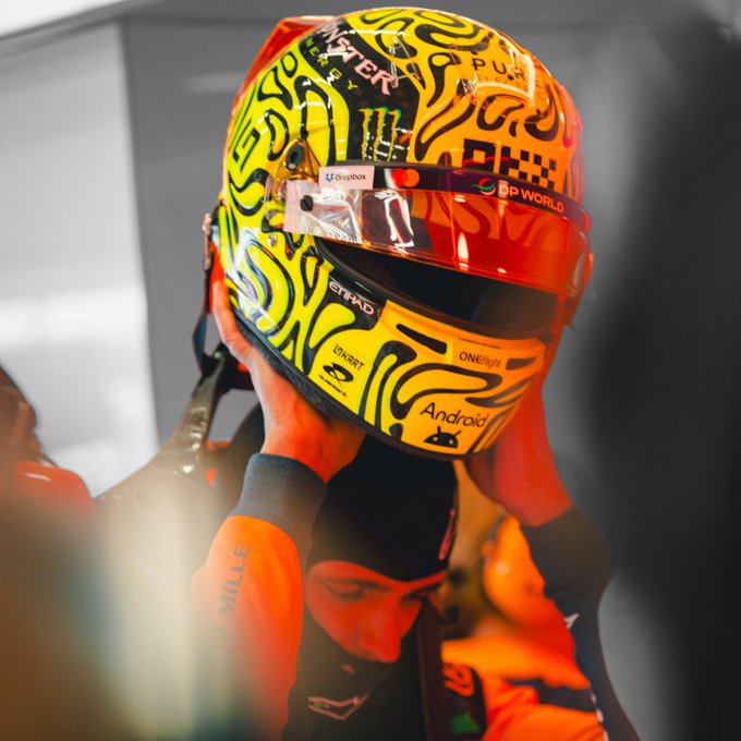 A Formula 1 driver puts on a vibrant orange and yellow helmet adorned with sponsors like Monster, Android, Rolex, and "DP World," preparing for the race. The image captures the focus and readiness behind the caption "Ready to go all in."