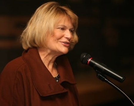 A blonde woman wearing a rust-colored jacket speaks into a microphone against a dark background.