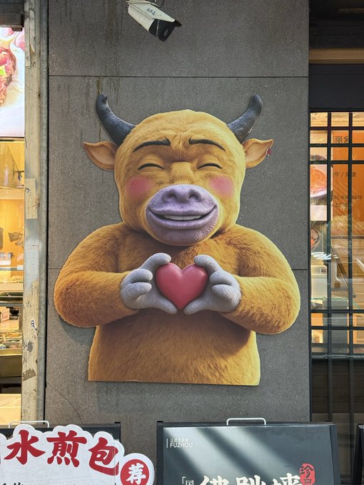 A whimsical 3D sculpture of a smiling brown bull making a heart shape with its hands is mounted on a gray pillar outside a shop with Chinese signage.