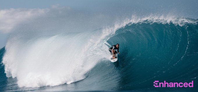 A surfer carves through a massive, curling wave while riding inside a frothy barrel of water. The "Enhanced" logo with its distinctive pink "E" emblem appears in the bottom right, tying the image to the post's shoutout to @enhanced_defi.